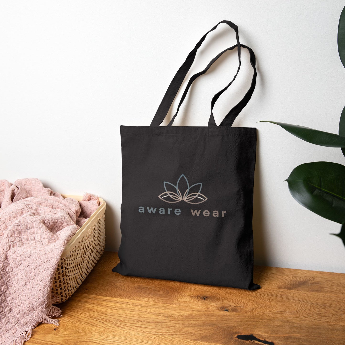 Black tote bag with 'aware wear' logo on a wooden surface with a plant in the background front view