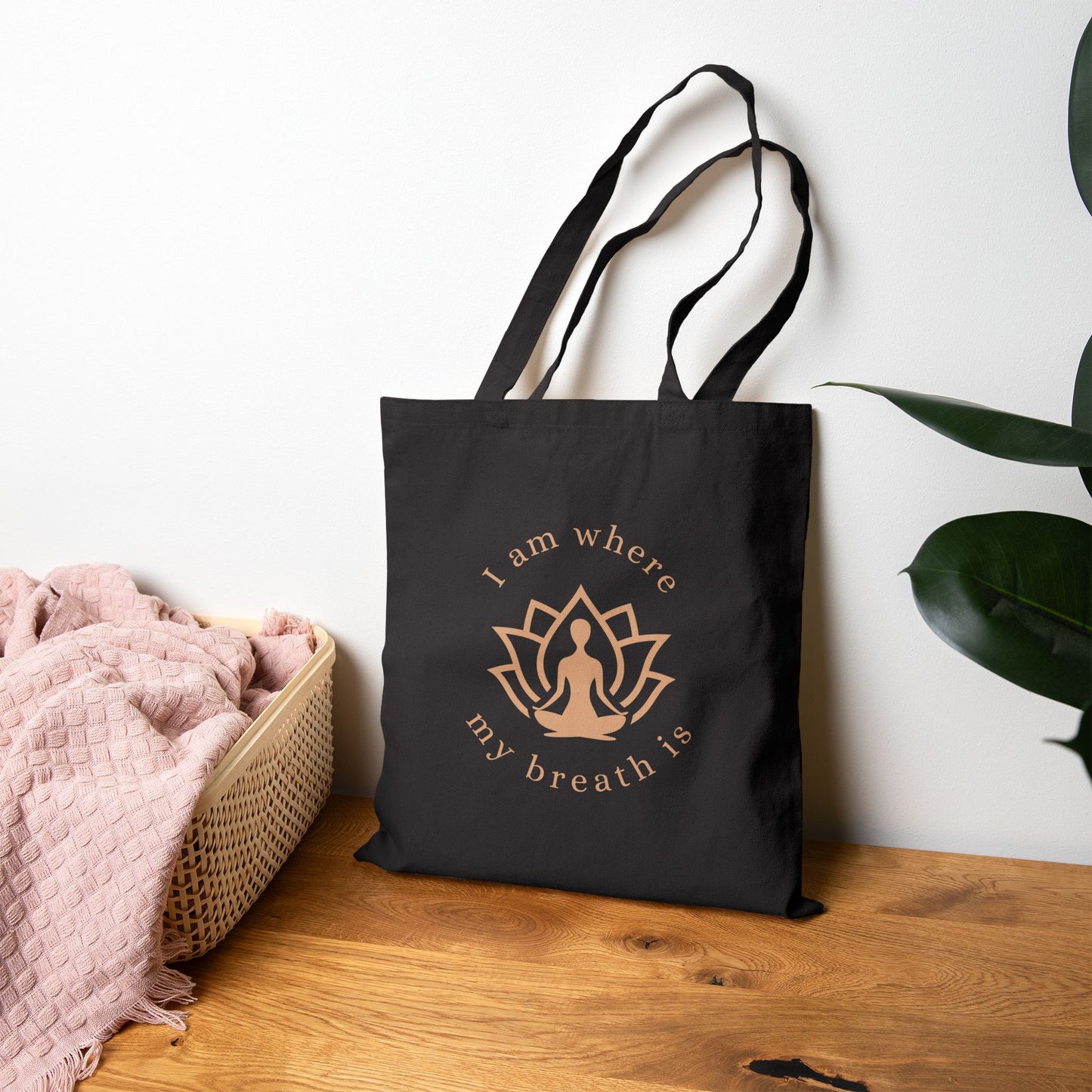 Black tote bag with a gold design and text on a wooden surface with a plant in the background