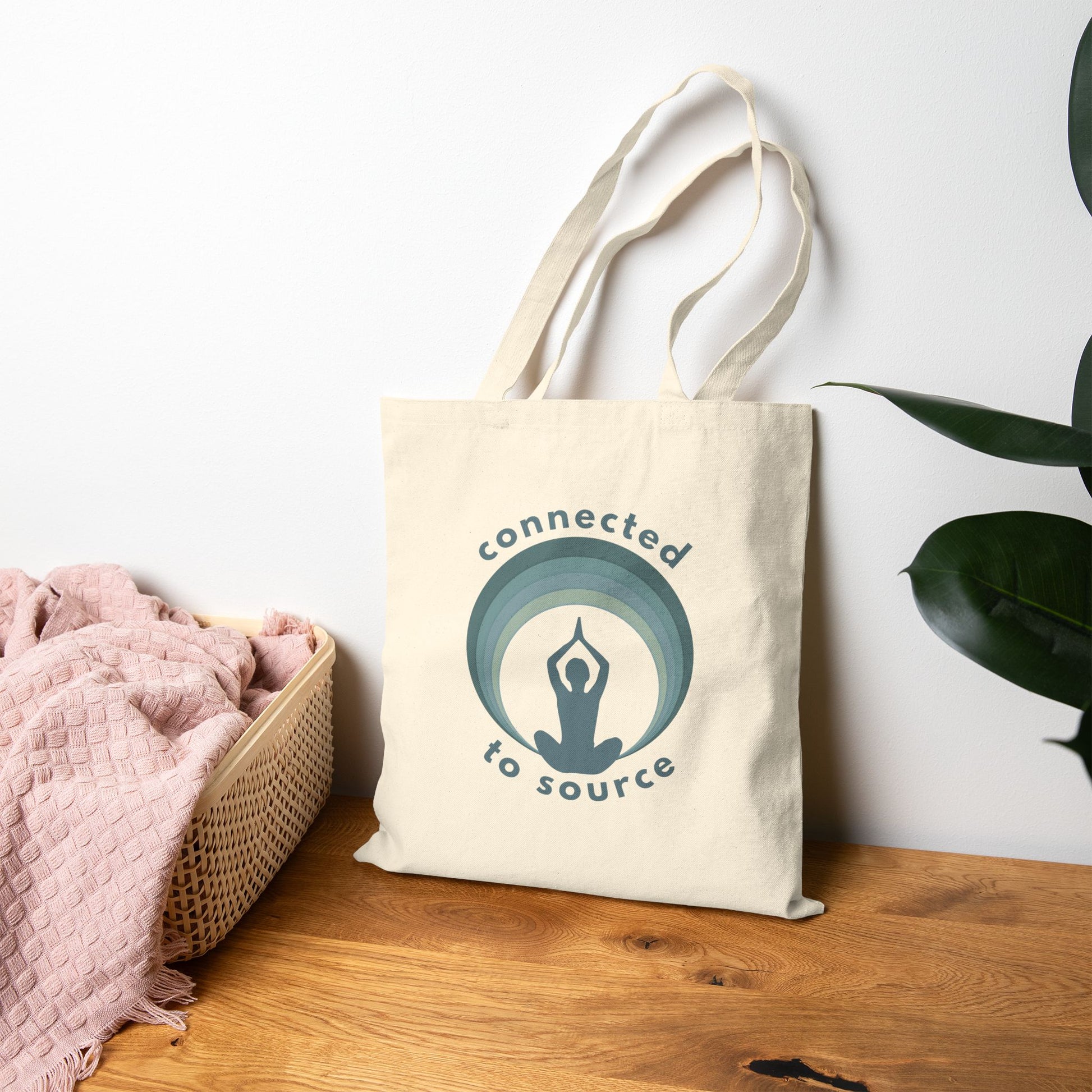 Tote bag with 'connected to source' logo on a wooden surface with a plant in the background