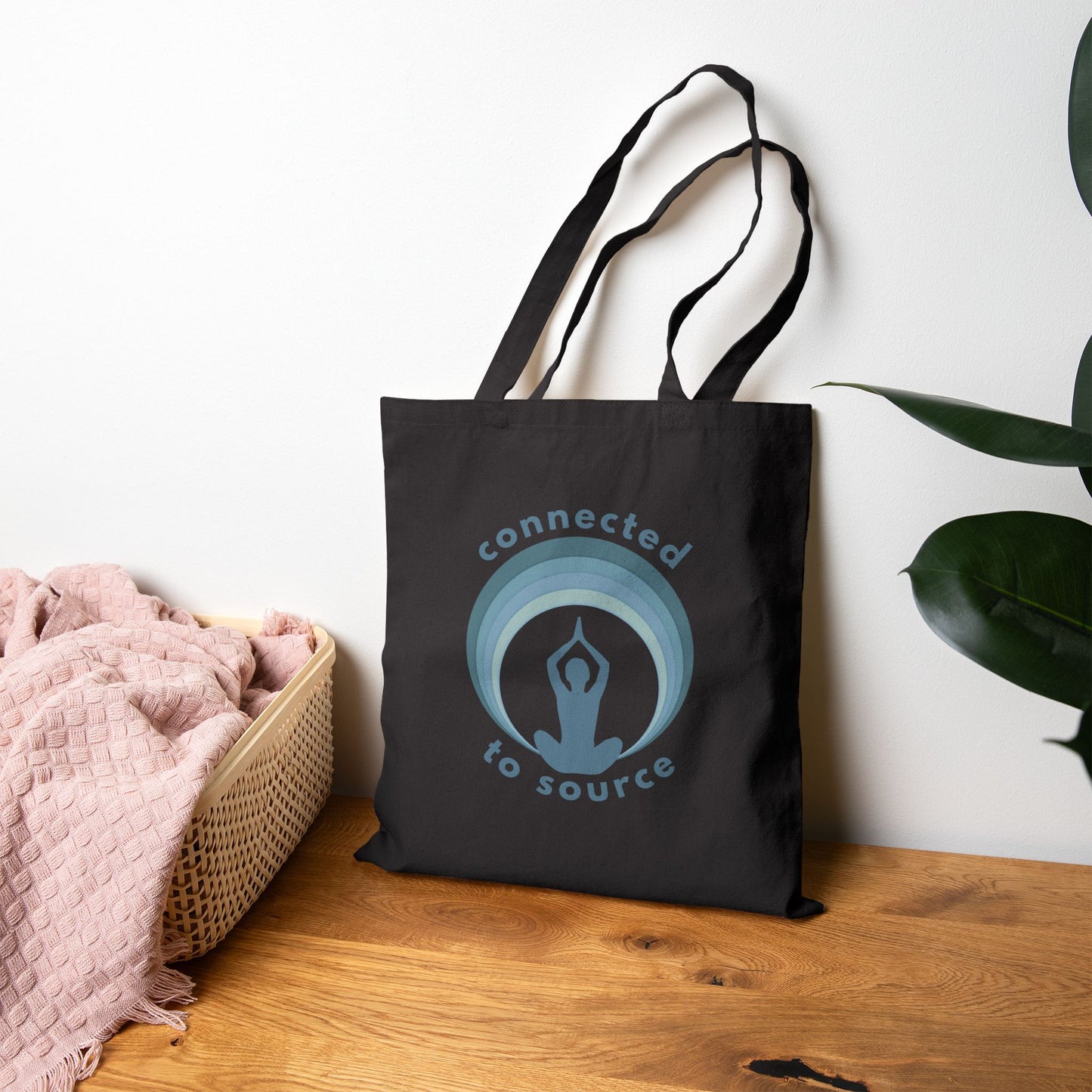 Black tote bag with 'connected to source' logo on a wooden surface with a plant in the background