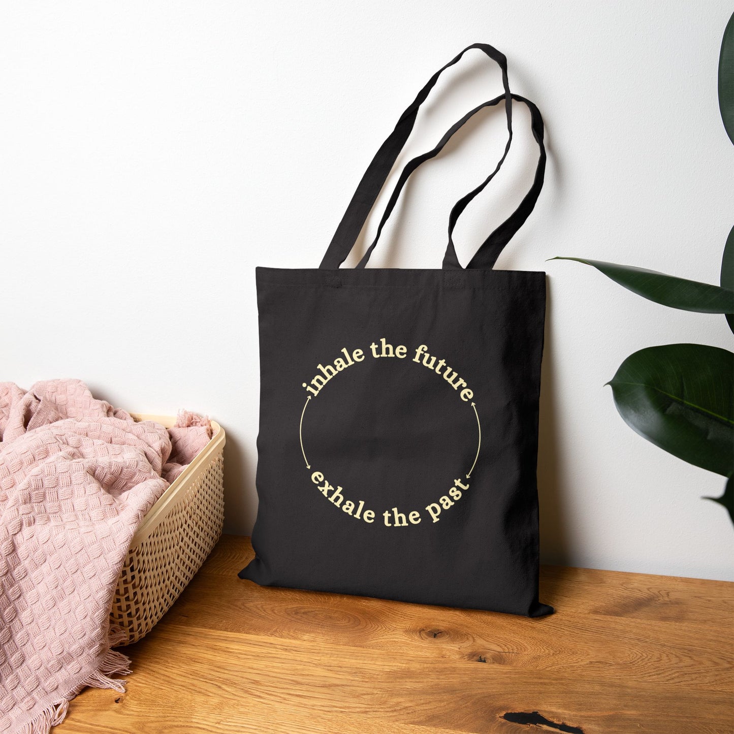 Black tote bag with text on a wooden surface with a plant in the background